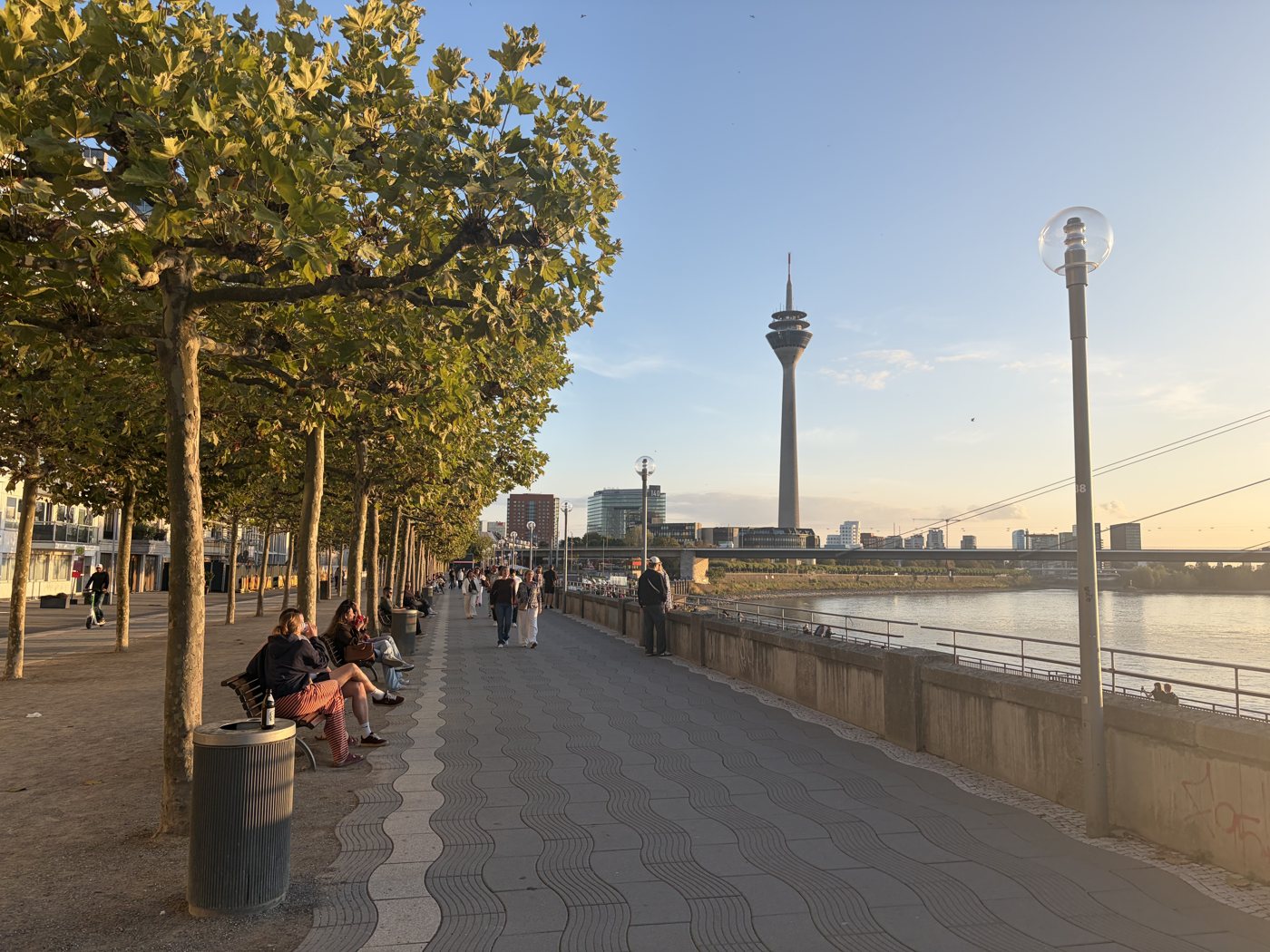 Menschen sitzen an der Düsseldorfer Rheinuferpromenade, im Hintergrund der Rheinturm und die Rheinkniebrücke im Abendlicht.